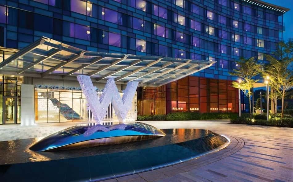 Hotel moderno com fachada de vidro azul, cobertura de vidro na entrada e fonte em forma de W.