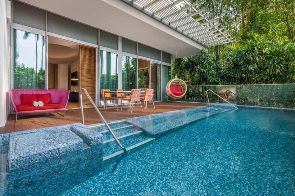 Hotel com piscina azul, deck de madeira, sofá rosa, poltronas laranja e cadeira suspensa vermelha, jardim tropical.