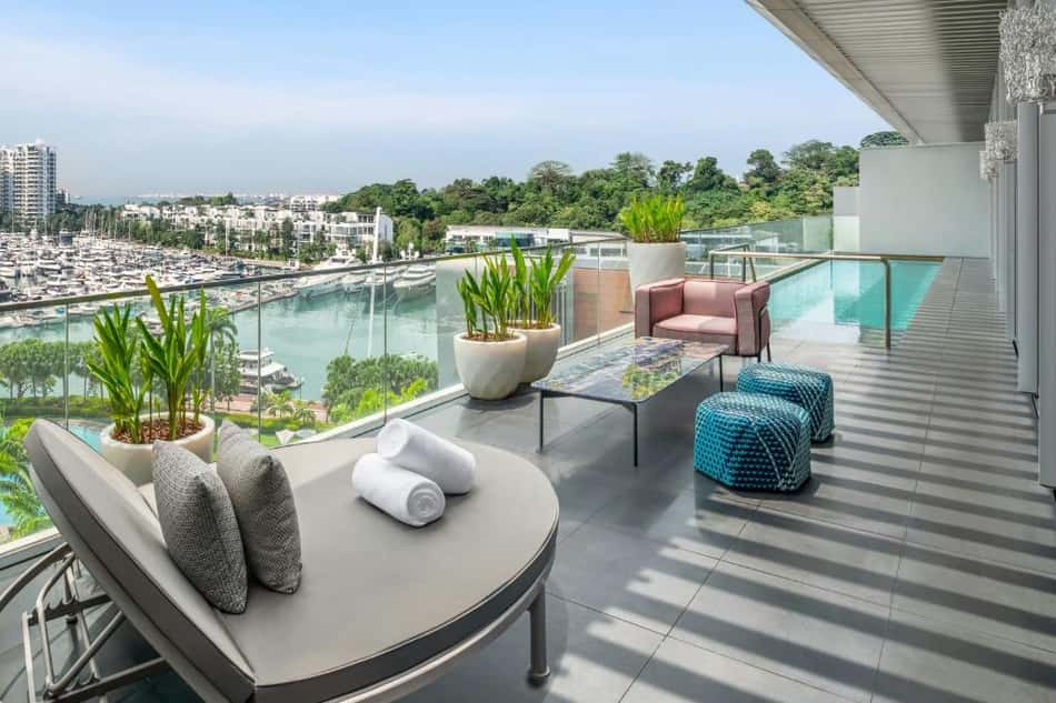 Varanda de hotel com piscina, mobiliário de lounge, plantas e vista para marina e cidade ao fundo.