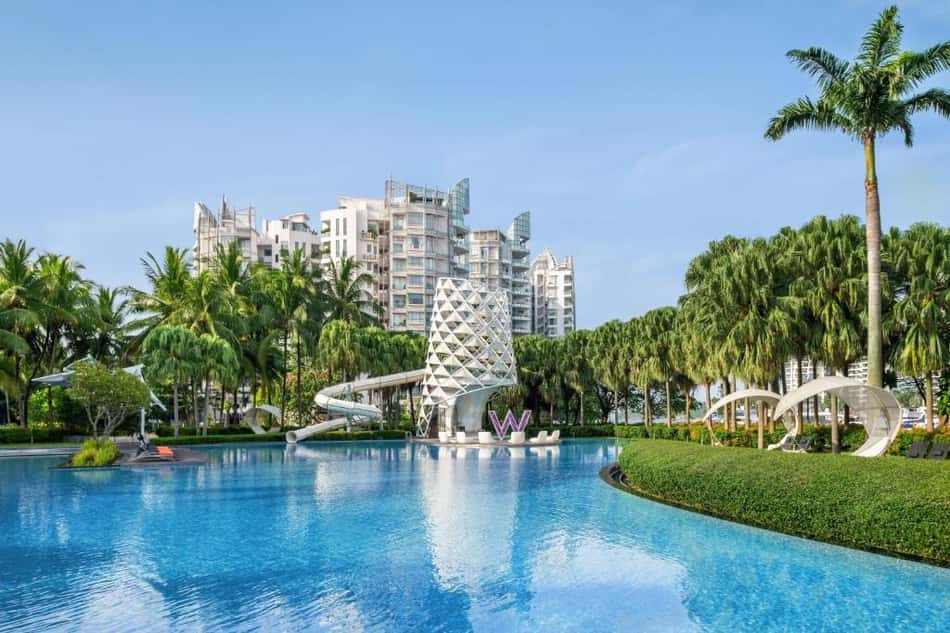 Resort com piscina azul cercada de palmeiras, torre escultórica branca ao centro e edifícios modernos ao fundo.