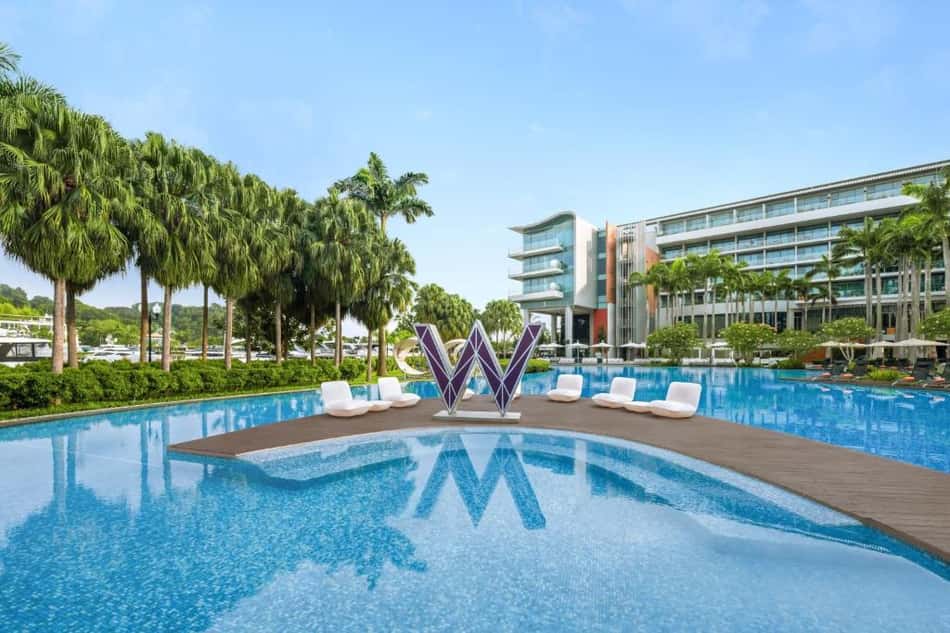 Piscina de hotel com ilha central, escultura em forma de W, palmeiras ao redor e prédio moderno ao fundo.