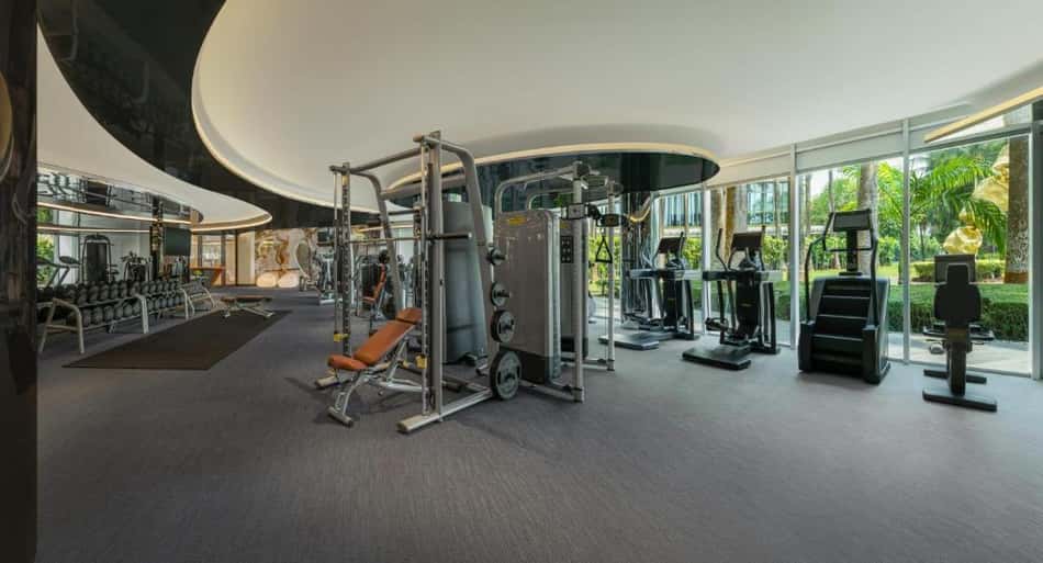 Academia de hotel com equipamentos de musculação e cardio, bancos, esteiras e janela ampla com área externa verde.