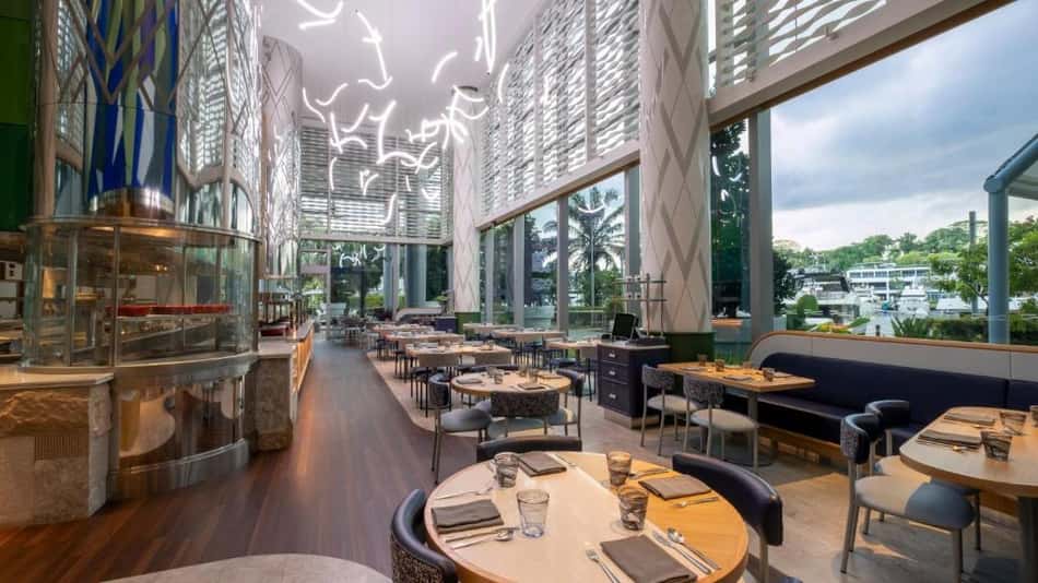 Restaurante de hotel com mesas postas, cadeiras azuis e janelas amplas, vista para o lago ao fundo.