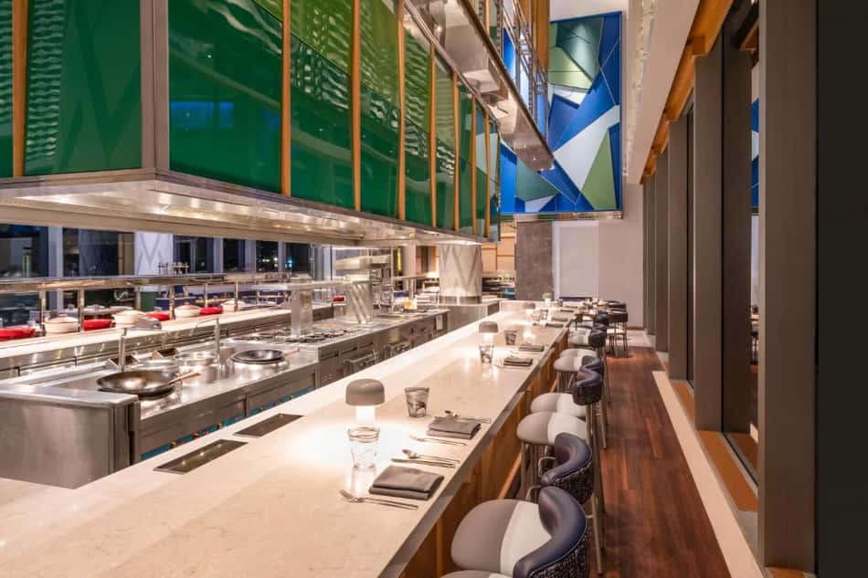 Cozinha aberta de hotel com balcão longo, cadeiras altas, bancada de mármore, aço inox e iluminação elegante.