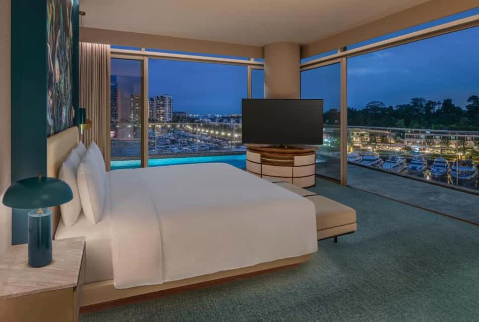 Quarto de hotel moderno com cama king, TV, bancada ao lado e varanda com vista para marina iluminada.