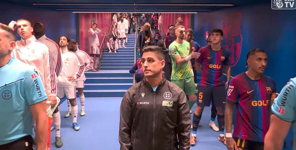 Grupo de jogadores de futebol no túnel, alguns com camisas listradas azul e bordô e um homem ao centro com casaco preto.