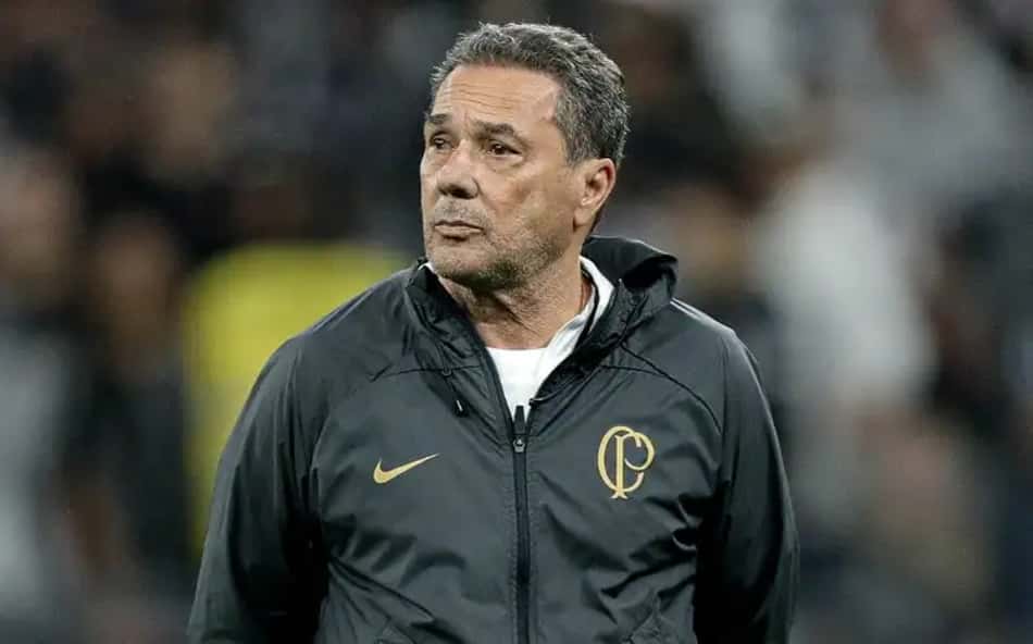 Treinador de futebol em casaco escuro, em campo, olhando para o lado.
