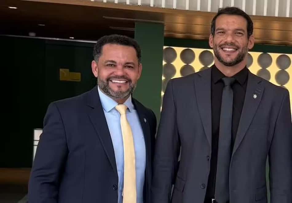 Dois homens de terno posam juntos, sorrindo, em ambiente interno.