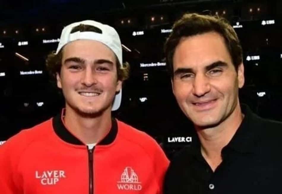 Dois homens em quadra de tênis; um com jaqueta vermelha Layer Cup World e outro com polo preto, cercados por banners.