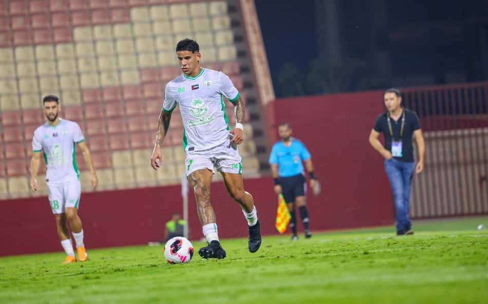 Jogador com uniforme branco e verde driblando a bola em campo, árbitro ao fundo e companheiros ao redor