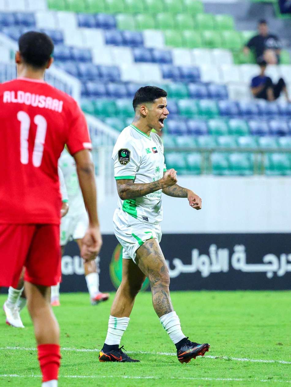 João Vítor Emirados, jogador com uniforme branco e verde, celebra em campo durante partida de futebol contra adversário de vermelho.
