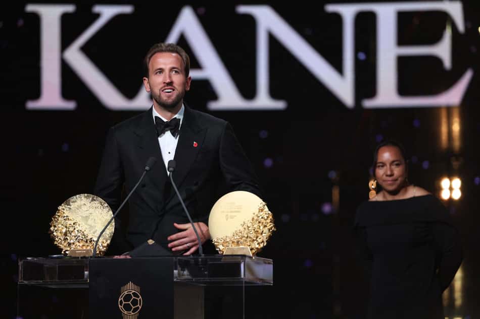 Homem em smoking no palco da premiação Kane, com dois troféus dourados, mulher de preto ao fundo.