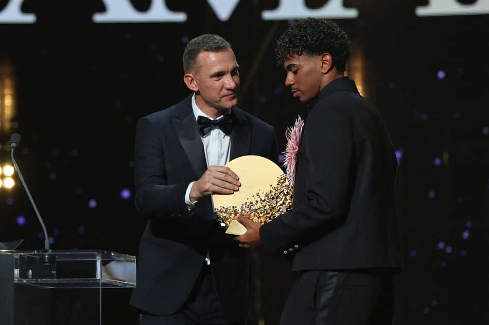 Lamine Yamal recebe prêmio em cerimônia esportiva, ao lado de apresentador, segurando troféu circular dourado com confetes.