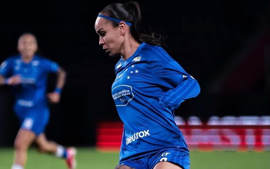 Letícia Cruzeiro em campo, jogadora de futebol, com uniforme azul e tatuagem na perna, dominando a bola.