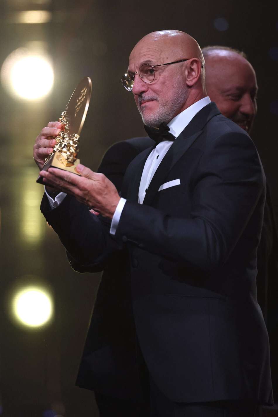 Homem de terno escuro com óculos segurando troféu dourado em cerimônia de premiação.