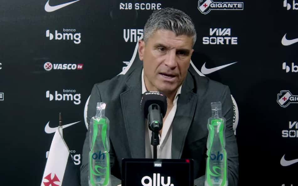 Treinador em coletiva de imprensa Vasco x Corinthians, diante de um microfone, com garrafas de água e backdrop com logos