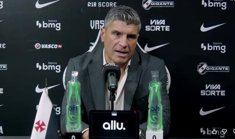 Treinador em coletiva de imprensa Vasco x Corinthians, diante de um microfone, com garrafas de água e backdrop com logos