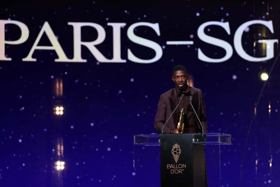 Homem em terno segura o troféu Ballon d'Or no palco, com fundo exibindo PARIS-SG.