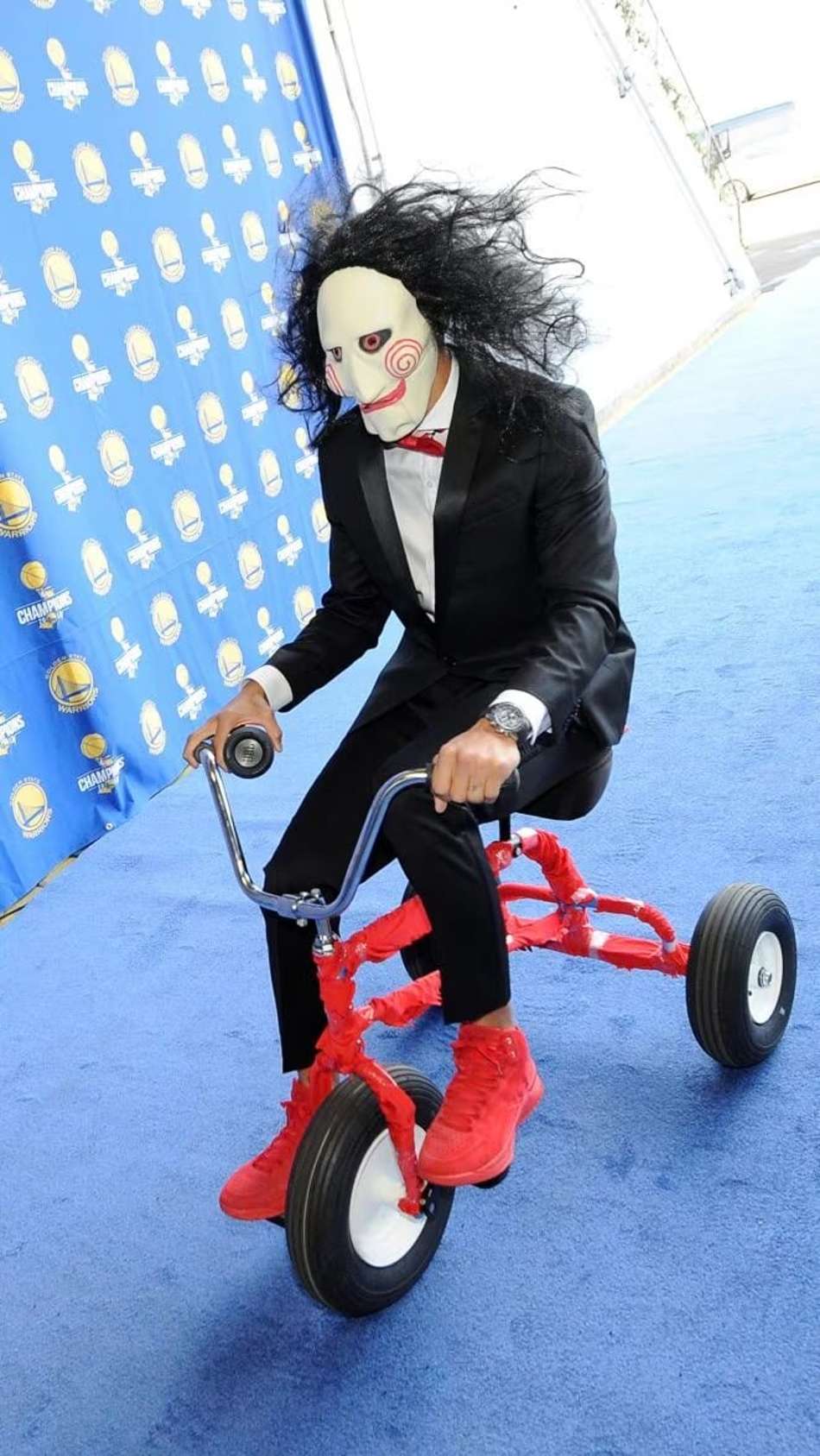 Participante em traje de Halloween, máscara de Jigsaw, pedalando triciclo vermelho; fundo azul com logotipos de basquete.