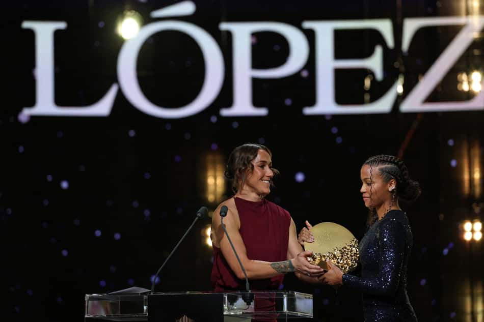 Apresentadora em vestido vinho entrega prêmio a jovem participante no palco, com letreiro Lopez ao fundo.