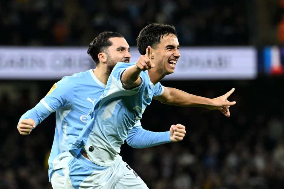 Dois jogadores do Manchester City comemoram; o da frente aponta, sorrindo, enquanto o outro vem atrás.