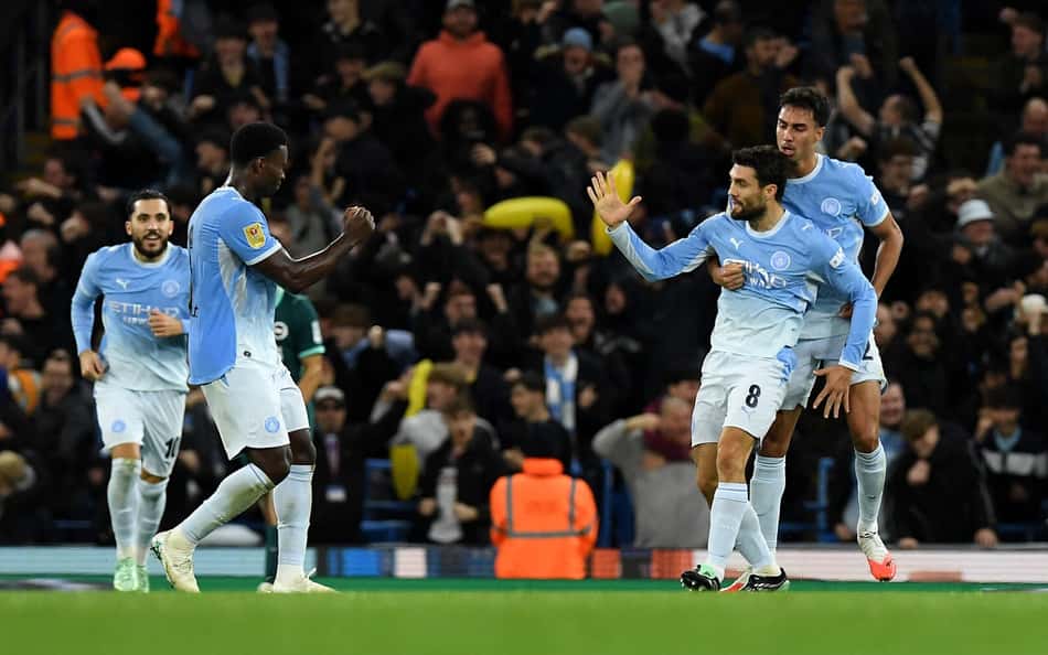 Jogadores do Manchester City comemoram gol durante partida, torcida ao fundo.