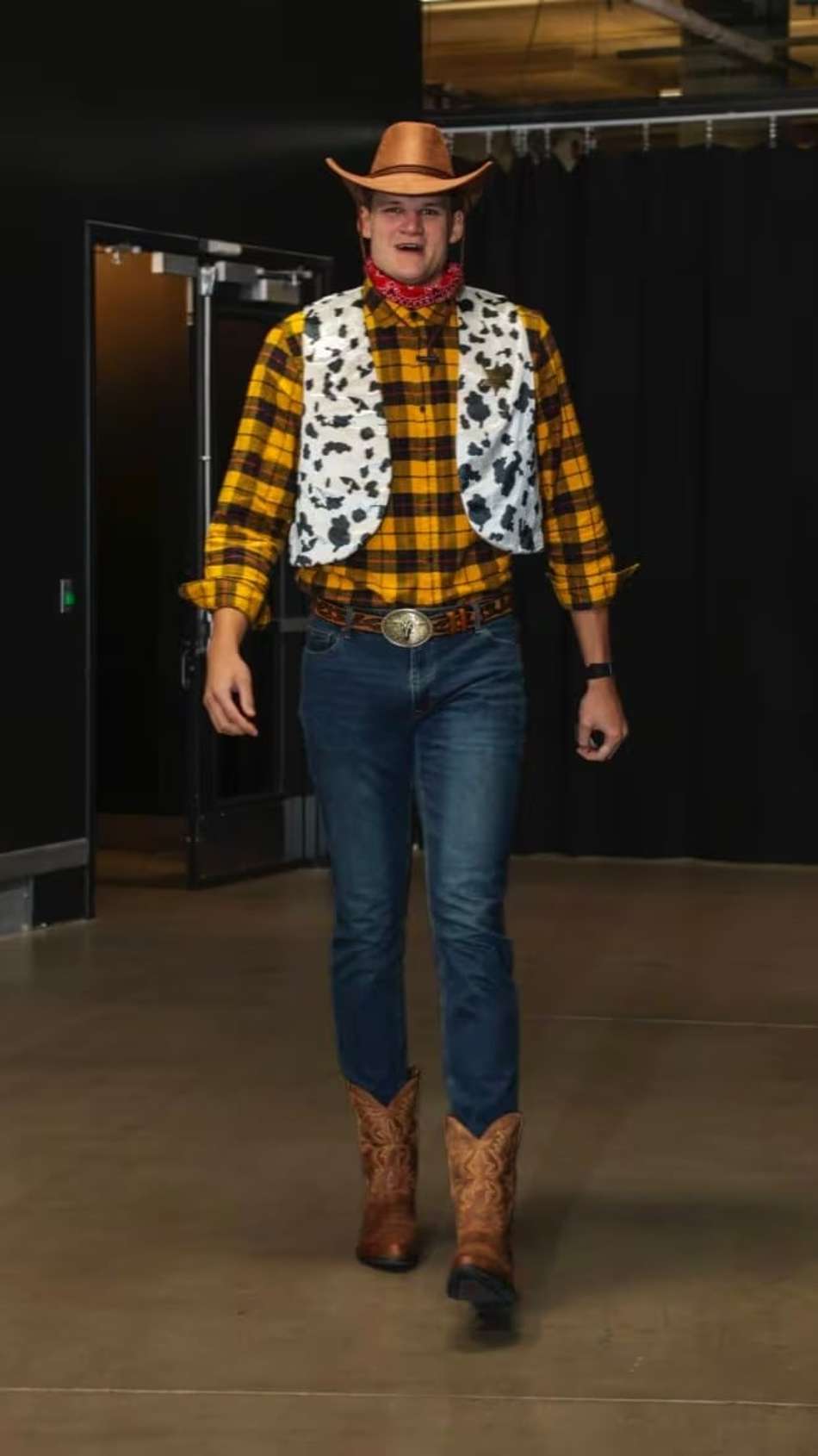 Indivíduo fantasiado de caubói Halloween, chapéu marrom, colete com estampa de vaca, camisa xadrez amarela, jeans e botas.