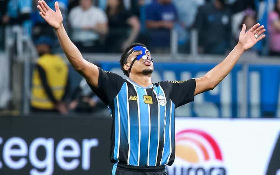 Wallace, jogador do Grêmio, celebra de joelhos com os braços erguidos em campo.