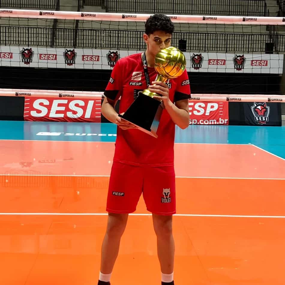 William Pereira, campeão paulista sub-21 de vôlei pelo Sesi/Bauru, segurando o troféu no piso da quadra.