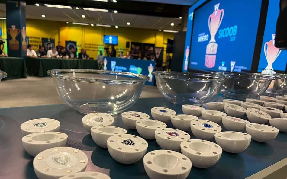 Mesa com várias tigelas brancas com emblemas de times; telão azul exibe troféu ao fundo.