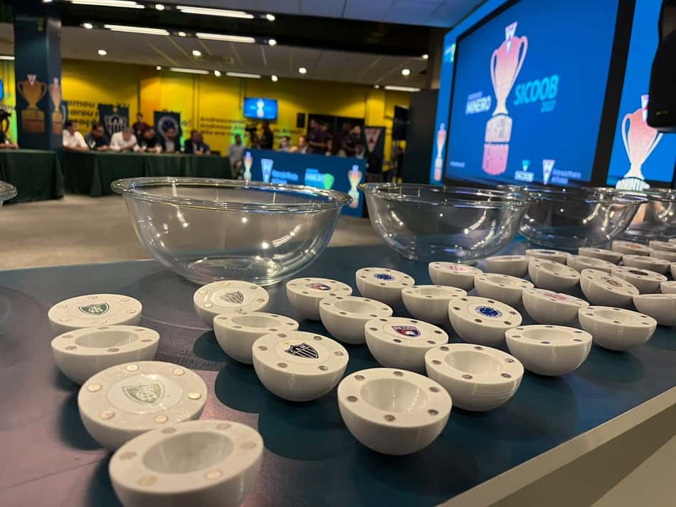 Mesa com várias tigelas brancas com emblemas de times; telão azul exibe troféu ao fundo.