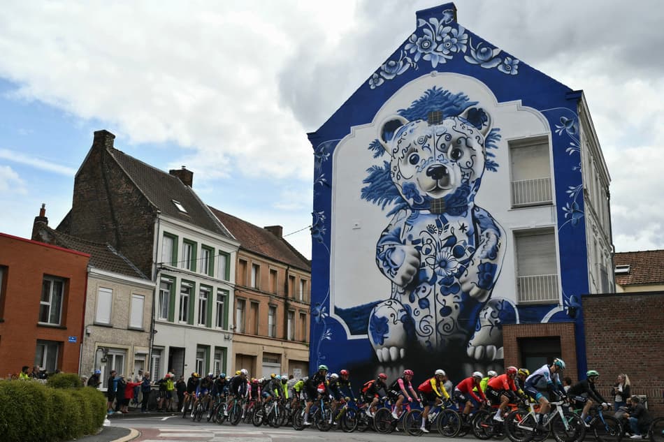 os ciclistas passam por um mural apelidado de 'Douceur d'Amand', na lateral de um prédio em Saint-Amand-les-Eaux (foto: Marco BERTORELLO / AFP)