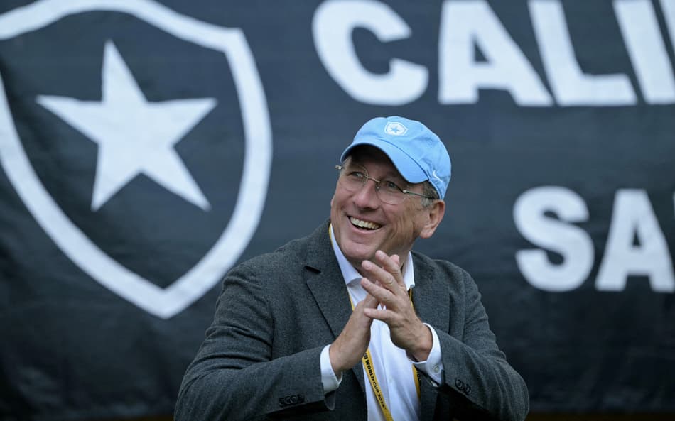 John Textor sorri e aplaude usando um boné azul em frente a um bandeirão do Botafogo no fundo (Foto: Juan Mabromata/AFP)