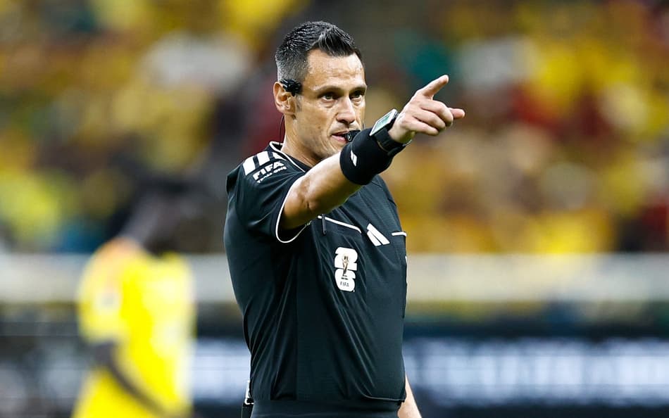 Maurizio Mariani, com camisa preta e com o emblema da Fifa, aponta com a mão direita para sinalizar marcação em Colômbia x RD Congo