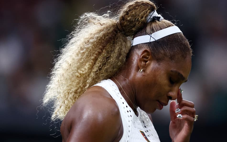 De faixa branca no cabelo e top branco, Serena Williams leva o dedo indicador esquerdo ao rosto em partida de Wimbledon