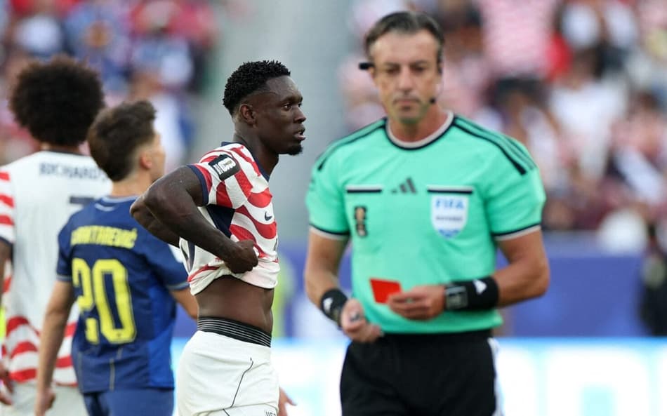 O árbitro Raphael Claus, em primeiro plano, segura um cartão vermelho; atrás dele, Balogun, o jogador expulso, levanta a camisa parcialmente e demonstra tristeza