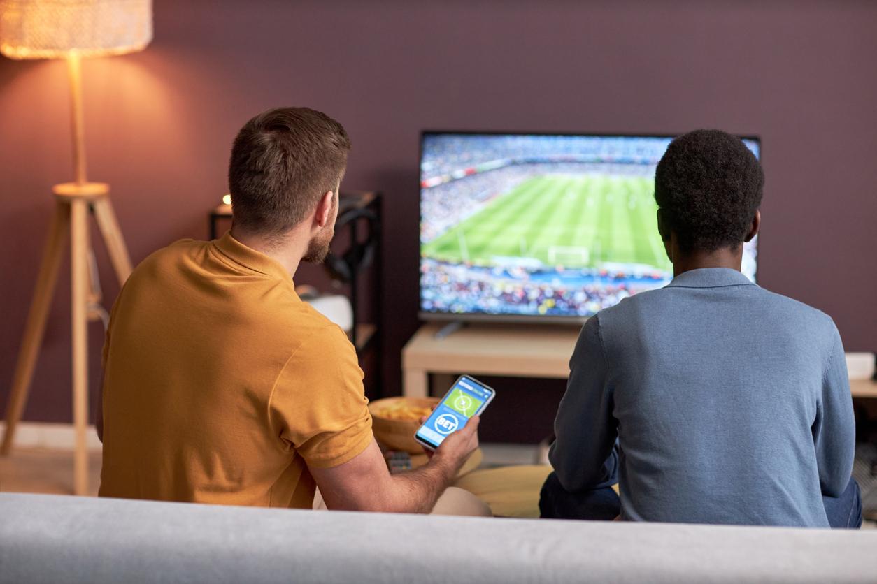 Two men watching football match on TV and holding smartphone with online bets
