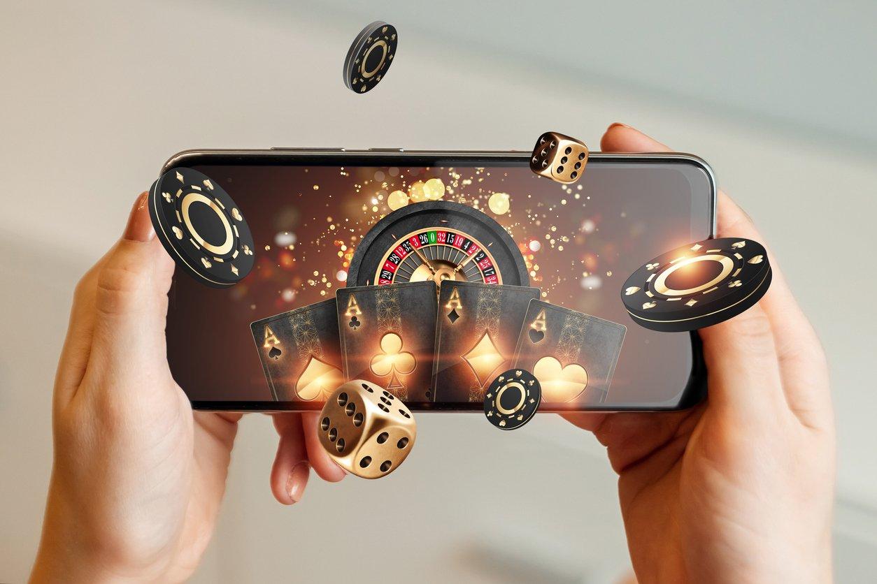 Creative background, online casino, in a man’s hand a smartphone with playing cards, roulette and chips, black-gold background. Internet gambling concept. Copy space